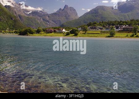Klares Wasser mit einem Dorf dahinter vor der Kulisse von Bergen unter einem blauen Himmel, Romsdalshornet, Raumadalen, Trollvegen, Andalsnes, Norwegen Stockfoto