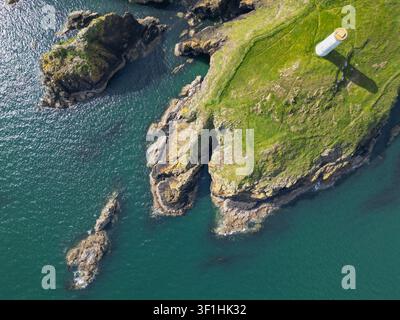 Luftaufnahme eines weißen Leuchtturms, der auf einer grasbewachsenen Klippenkante steht und über das tiefe blaue Meer mit felsigen Ausläufern und zerklüfteter Küste in Waterford blickt Stockfoto
