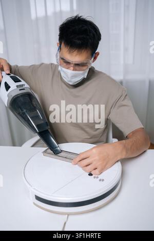 Vertikales Porträt eines jungen Mannes, der eine Schutzmaske und eine Schutzbrille trägt und einen schmutzigen Filter von einem Roboterstaubsauger mit einem kleinen Handstaubsauger reinigt Stockfoto