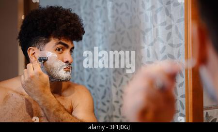 Junger Mann, der seinen lockigen Bart mit einem Rasierer und Schaum pflegt Stockfoto
