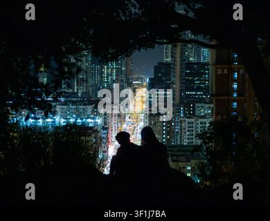 Eine Silhouette eines jungen Paares, das nachts auf einem Hügel unter einem Baum sitzt und die schimmernde blaue Stadtlandschaft dahinter betrachtet, Hongkong Stockfoto