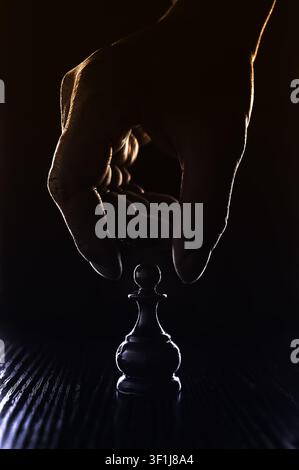 Ein dramatisch kantenbeleuchtetes, farblich abgestuftes Foto einer Hand, die einen Schachbauern bewegt, mit einem dunklen Hintergrund, der innovative Planung im Geschäftsleben darstellt. Stockfoto