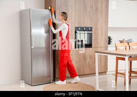 Junge Frau in roten Latzhosen und orangefarbenen Handschuhen misst einen Kühlschrank in einer modernen Küche für Heimwerkerarbeiten Stockfoto