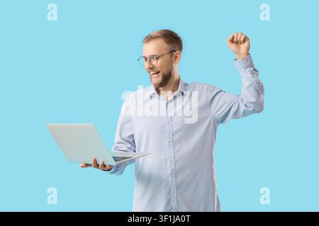 Begeisterter junger Mann mit Bart und Brille, der einen Laptop hält, feiert den Erfolg mit einer erhobenen Faust vor einem blauen Hintergrund Stockfoto