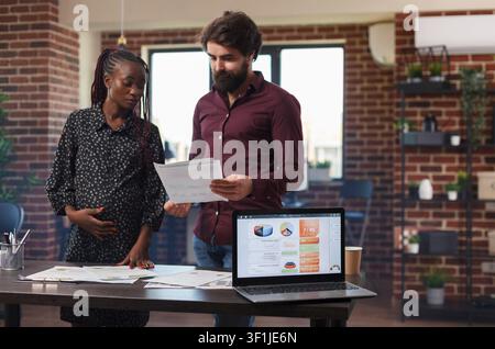 Laptop zeigt Geschäftsanalyse-Diagramme, während eine schwangere Geschäftsfrau und ein Kollege Unternehmensumsatz und Buchhaltungsdaten in einem Büro analysieren. Stockfoto