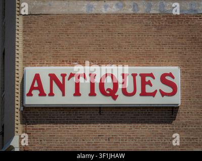 Großes weißes Werbeschild mit fetten roten Buchstaben, die „Antiques“ lesen, montiert an einer alten verwitterten roten Backsteinfassade im Sonnenlicht Stockfoto