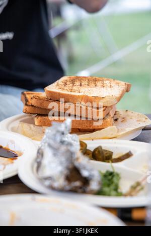Scheiben gegrillten Toasts sind auf einem Pappteller bei einem Außenevent gestapelt, wobei folienverpackte Speisen und Gewürze im Vordergrund auf einem Holztisch sichtbar sind. Stockfoto