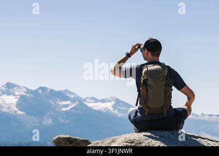Ein Wanderer, der auf einem Bergausblick sitzt, richtet seine Mütze, während er in die schneebedeckten Gipfel in der Ferne blickt. Stockfoto