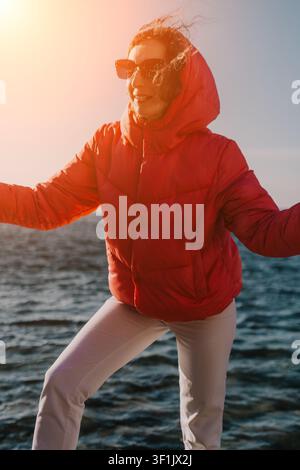 Frau in roter Jacke am Strand bei Sonnenuntergang, glückliche junge Frau, die warme Winterkleidung trägt und während der goldenen Stunde am Meer steht Stockfoto