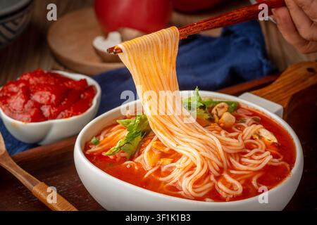 Traditionelle chinesische Tomatensuppen-Nudeln in einer weißen Schüssel mit Holzgriff, garniert mit Pilzen und Grünzeug, serviert auf einem dunklen Holztray. Stockfoto