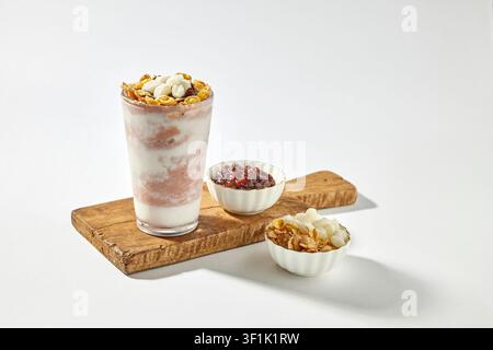 Granola-Yoghurt-Parfait-Getränk in einem hohen Glas, serviert auf einem Holztablett mit Schüsseln mit Belägen wie roten Bohnen, Cornflakes und Nüssen auf einem hellgrauen Hintergrund Stockfoto