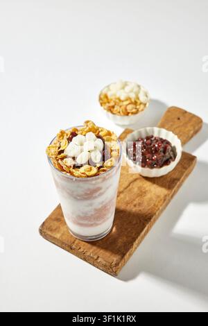 Granola-Yoghurt-Parfait-Getränk in einem hohen Glas, serviert auf einem Holztablett mit Schüsseln mit Belägen wie roten Bohnen, Cornflakes und Nüssen auf einem hellgrauen Hintergrund Stockfoto