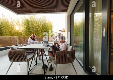 Familie entspannt sich auf modernem Hausbalkon mit Laptop und Getränken während einer Sonnenuntergangsterrassenparty Stockfoto