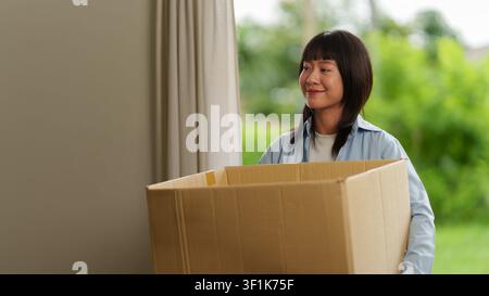 Lächelnde asiatische Frau trägt während des Umzugs einen Karton Stockfoto