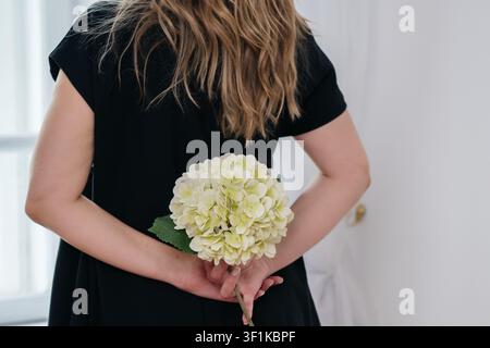 Eine Rückansicht einer Frau mit langen blonden Haaren, die ein schwarzes Kleid trägt und einen einzelnen Stiel einer blühenden weißen Hortensienblüte hält und verbirgt Stockfoto