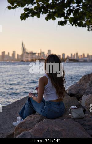 Junge Frau sitzt auf Felsen und genießt den Blick auf die Skyline unter Baumästen Stockfoto