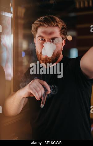 Junger bärtiger Mann, der Rauch ausatmet, während er eine Zigarre hält, in einem stimmungsvollen Innenporträt Stockfoto