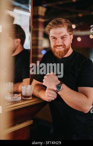 Lächelnder bärtiger Mann an einer Bar, der sich mit einem Getränk und einer Uhr auf der Theke lehnt, Freunde zusammen Stockfoto