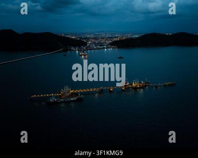 Beleuchtetes Ölterminal und Frachtschiffe, die während der Blauen Stunde an einem Industriepier in Si Racha Chon Buri, Thailand, festgemacht sind. Stockfoto