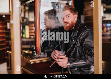 Bärtiger Mann in Lederjacke raucht eine Zigarre und hält an der Theke einer Bar ein Getränk Stockfoto