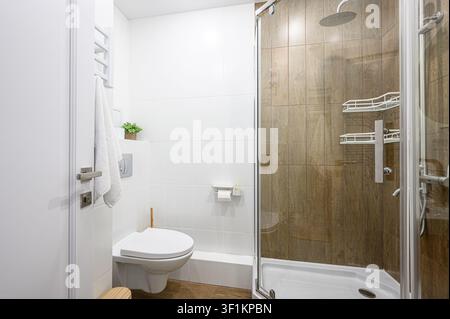 Helles Badezimmer mit einer Glasdusche mit Holzoptik-Fliesen, weißen Wänden, Toilette und Handtuchhalter. Sauberes und modernes Design Stockfoto