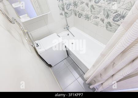 Oberblick auf ein helles Badezimmer mit einer Waschmaschine neben einer weißen Badewanne, grauen Fliesenböden und blattgemusterten Fliesen an der Wand. Eine Show Stockfoto