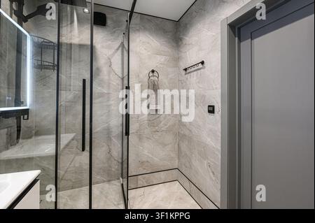 Modernes Badezimmer mit einer Glasdusche mit schwarzen Armaturen und marmor gemusterten Fliesen. Rechts ist eine graue Tür zu sehen. Sauberes, minimalistisches Design Stockfoto