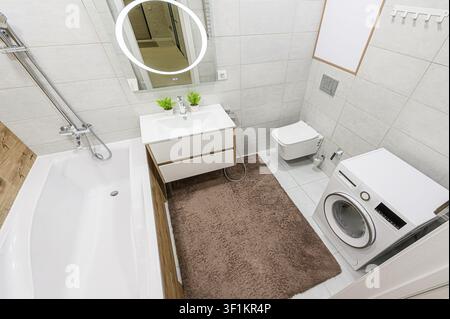 gut beleuchtetes, modernes Badezimmer mit weißen und grauen Fliesen, einer Badewanne, einer Toilette, einem Waschbecken mit beleuchtetem Spiegel, einer Waschmaschine und einem braunen Teppich Stockfoto
