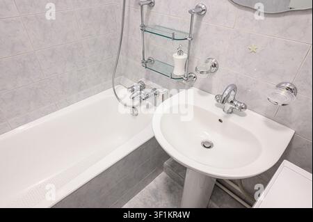 Sauberes Badezimmer mit einer weißen Badewanne, einem Standwaschbecken und gefliesten Wänden. Chrom-Armaturen und Glasregale verleihen einen Hauch von Modernität Stockfoto