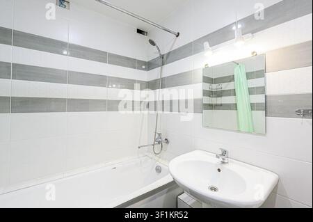 Sauberes Badezimmer mit einer weißen Wanne, einem Waschbecken, gefliesten Wänden mit grauen Streifen und einem Spiegel mit Licht Stockfoto