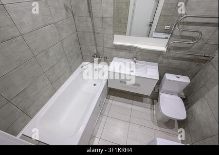 Sauberes, modernes Badezimmer mit grauen Fliesen, einer weißen Badewanne, Waschbecken, Toilette und einem beheizten Handtuchhalter Stockfoto