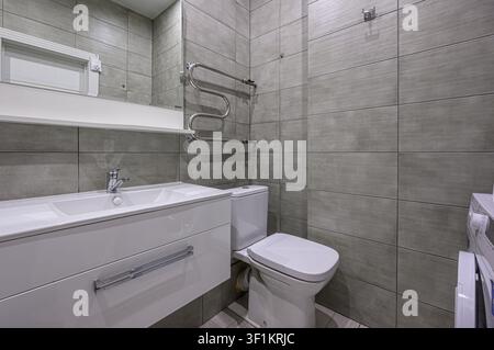 modernes Badezimmer mit grauen Fliesenwänden, einem weißen Waschtisch mit Waschbecken, einer Toilette und einem Handtuchheizkörper. Der Raum ist gut beleuchtet, was eine saubere und m Stockfoto