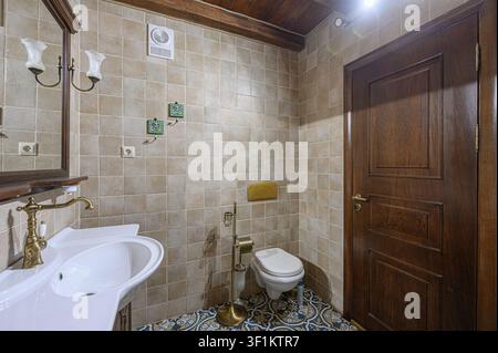 gemütliches Badezimmer mit beigen Fliesen, einem weißen Waschbecken und einer Holztür. Ein detaillierter gemusterter Boden verleiht Charme Stockfoto
