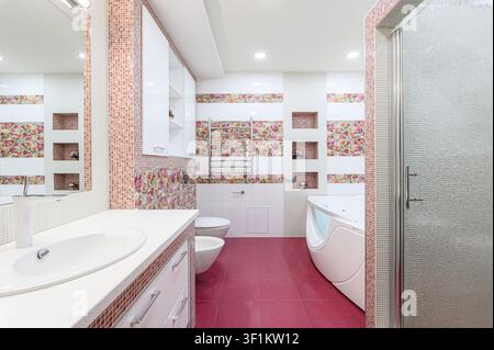 Helles Badezimmer mit pinkfarbenem Fliesenboden, weißen Schränken, floralen Wandakzenten, Dusche, Wanne, Toilette und Waschbecken Stockfoto