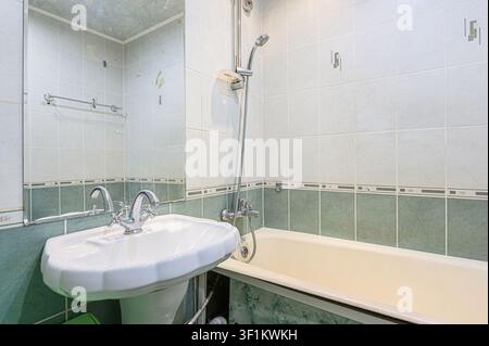 Badezimmer mit weißen und grünen Fliesen. Ein Waschtisch mit Chrom-Armaturen steht neben einer Badewanne mit Duschkopf. Ein Spiegel reflektiert einen Teil des Raumes Stockfoto