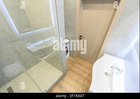 helles Badezimmer mit Glasdusche, modernem Waschbecken, einer Tür und einer Waschmaschine. Der Raum ist mit grauem marmorähnlichem Material gefliest und mit hellem Holz Stockfoto