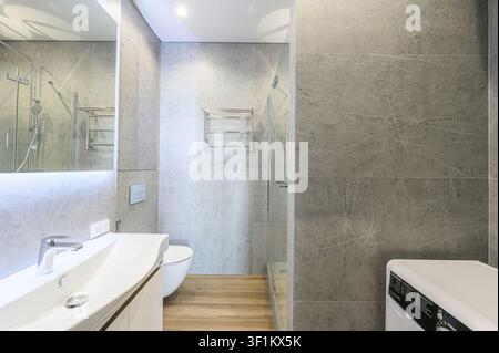 modernes Badezimmer mit grauen marmorartigen Fliesen, einem weißen Waschbecken und Toiletten, einem Spiegel mit eingebauter Beleuchtung und Holzoptik-Bodenbelag Stockfoto