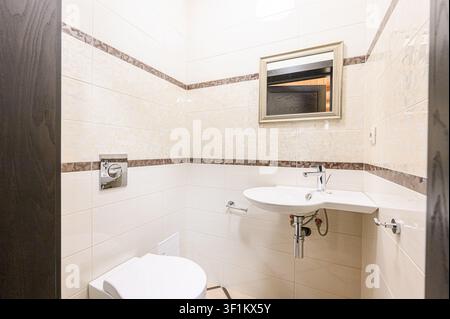 helles Badezimmer mit beige-tonigen Fliesen, einer weißen Toilette und einem Waschbecken. Ein Spiegel hängt an der Wand, und ein dunkler Holzrahmen des Türrahmens ist sichtbar Stockfoto