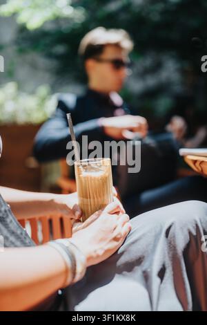Frau hält Eiskaffee in einem Straßencafé an einem sonnigen Tag mit verschwommenem Gast im Hintergrund Stockfoto