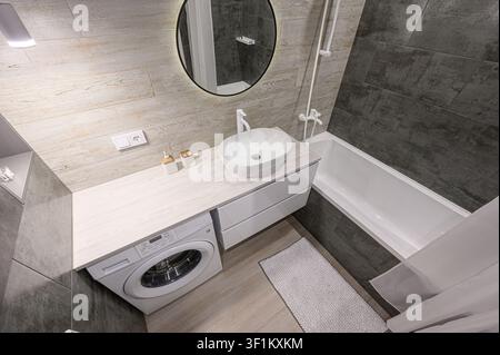 modernes Badezimmer mit hellen Fliesen in Holzoptik, einem weißen Waschbecken, einer Waschmaschine und einer Badewanne mit grauen Fliesen Stockfoto