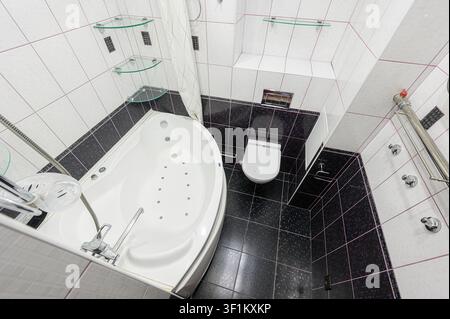 Ansicht von oben eines modernen Badezimmers mit einer Whirlpoolwanne, einer Toilette und gefliesten Wänden in Schwarz und Weiß. Glasregale verleihen einen Hauch von Eleganz Stockfoto