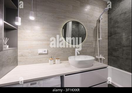 Modernes Badezimmer mit Holzoptik-Fliesen, einem runden beleuchteten Spiegel und schlanken weißen Armaturen. Sauberes, minimalistisches Design Stockfoto