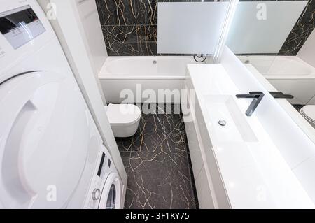 modernes Badezimmer mit weißen Armaturen, schwarzen und goldenen Marmorfliesen und einer gestapelten Wasch-/Trocknereinheit Stockfoto