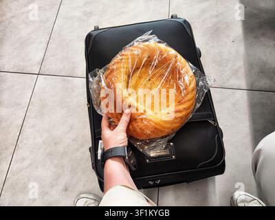 Eine Person hält einen runden Brotlaib, der in Plastik eingewickelt ist, während sie neben einem Koffer in einem Flughafenterminal sitzt. Stockfoto