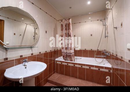 gut beleuchtetes Badezimmer mit braunen und beigen Fliesen, einem weißen Waschtisch, einem ovalen Spiegel und einer Badewanne mit einem gemusterten Vorhang Stockfoto