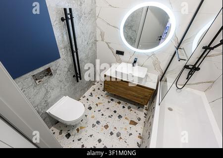 modernes Badezimmer mit Toilette, Waschbecken, rundem beleuchtetem Spiegel und Badewanne. Der Raum hat einen stilvollen Terrazzoboden und marmorierte Wände Stockfoto