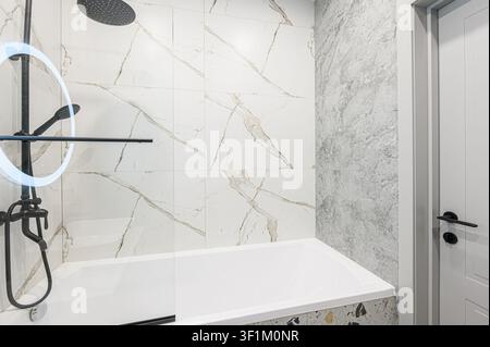 Modernes Badezimmer mit weißen Marmorfliesen, einem schwarzen Duschkopf, einer weißen Badewanne und einer grauen Marmorwand Stockfoto