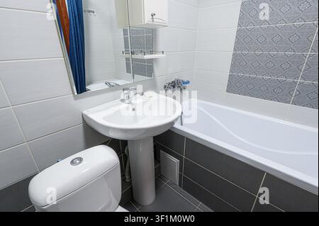sauberes Badezimmer mit weißen und grauen Fliesen, einer Toilette, einem Waschbecken, einer Wanne und einem Handtuchheizkörper Stockfoto