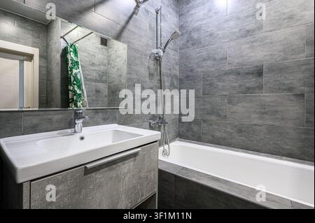 modernes Badezimmer mit grauen Fliesenwänden, einem weißen Waschtisch mit grauem Schrank, einem Spiegel und einer weißen Badewanne mit Chrom-Armaturen Stockfoto