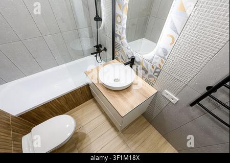 Modernes Badezimmer mit einem Aufsatzwaschbecken auf einem hellen Holzwaschtisch, einer Badewanne, gefliesten Wänden, einem gemusterten Spiegel und schwarzen Armaturen Stockfoto